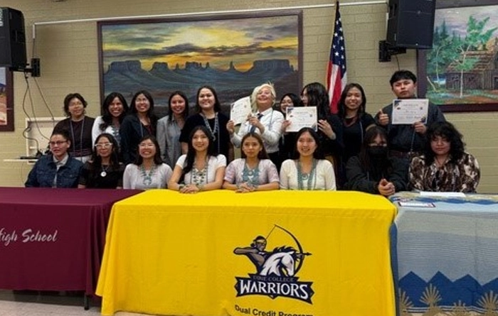 Two rows of students sit and stand behind a table and pose for a photo with a few of them holding up certificates.