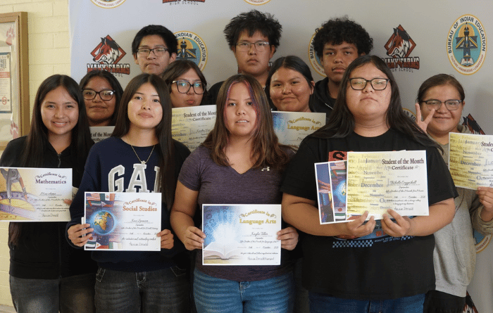 Students pose with certificates recognizing their academic achievements in front of a school backdrop.