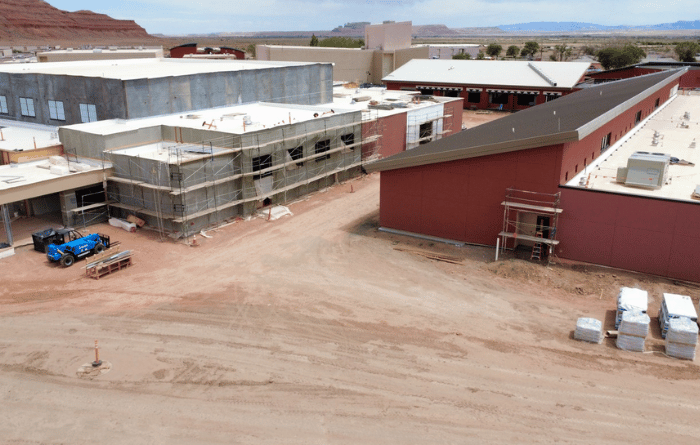 Aerial view of a school campus under construction, with red and gray buildings, scaffolding, and construction materials across the dirt-covered site.