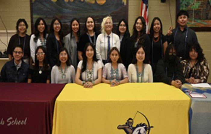 Group photo of approximately 20 students and staff standing and seated behind three tables with colored tablecloths and school logos, indoors. Most are smiling and wearing casual or semi-formal clothing; an American flag and artwork are visible in the background.
