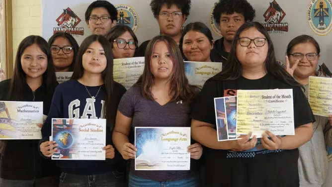 Students pose with certificates recognizing their academic achievements in front of a school backdrop.