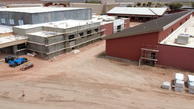 Aerial view of a school campus under construction, with red and gray buildings, scaffolding, and construction materials across the dirt-covered site.